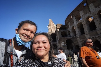 Picture of a man and woman standing in front of the Colosseum in Rome with people milling about underneath clear, blue skies.