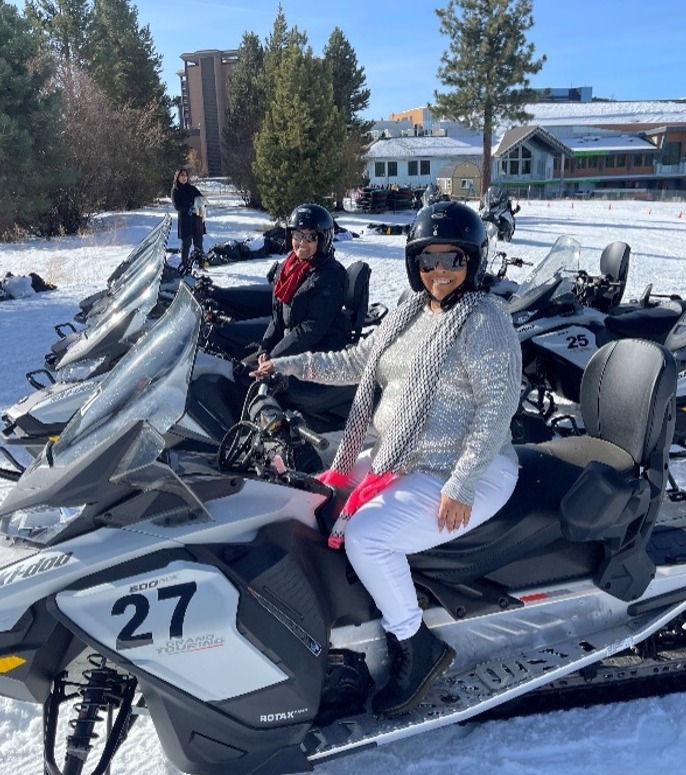 Two family members smiling, sitting on snowmobiles and surrounded by snow, evergreens and sunshine