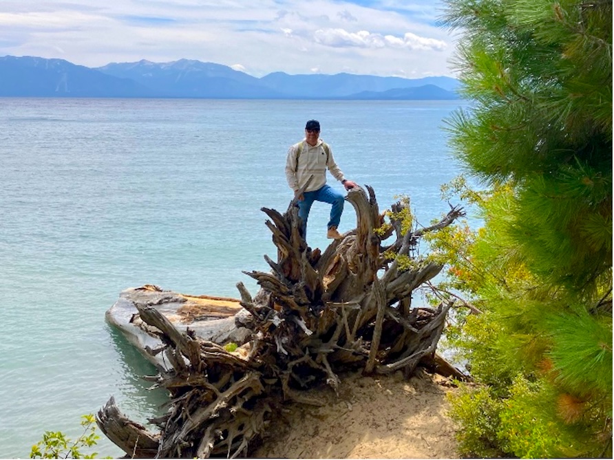 Man posing along a body of water, trees in foreground and mountain range in background.