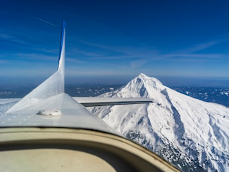 Airplane in flight with snow-capped mountaintops below, blue skies above.   