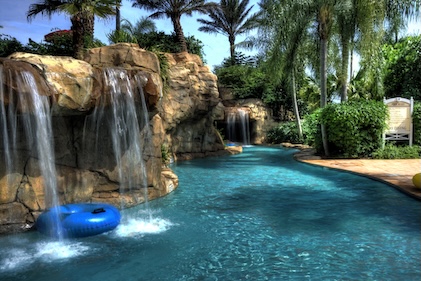 Mid shot of a lazy river with tall brown rocks and waterfalls on the left and palm trees in the distance.