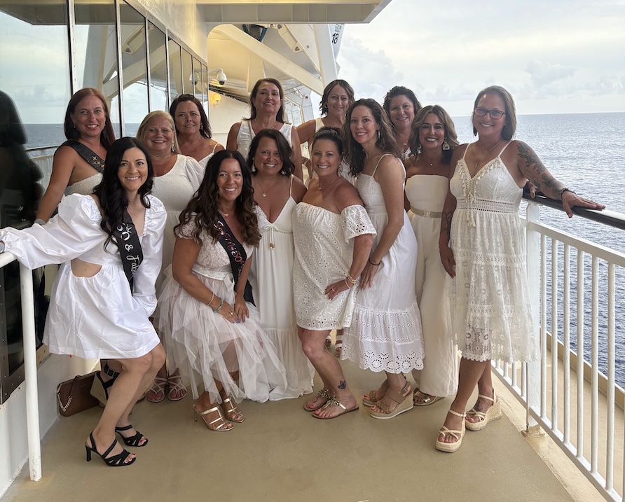 Group of 13 female friends in white dresses on a cruise deck.