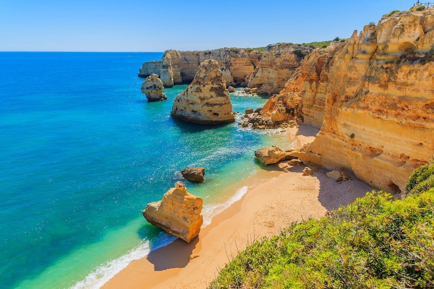 Beach scene in Portugal featuring bright blue water, rock formations and greenery.