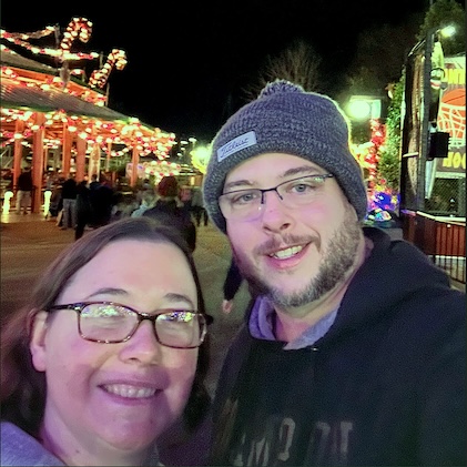 Couple enjoying a night out surrounded by buildings and attractions decorated in Christmas lights. 