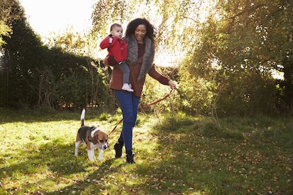 Woman holding a baby, both dressed in cool-weather clothing, walking a beagle while strolling through grass and surrounded by greenery