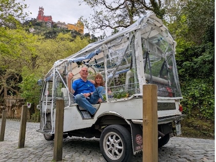 Couple aboard a white tuk tuk on Portugal's cobblestone streets, with leafy green trees in the background.