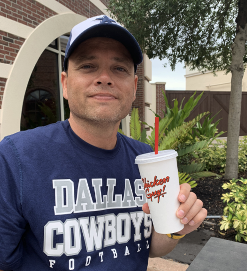 A smiling young man in a Dallas Cowboys football T-shirt and cap with a white cup and straw in his hand