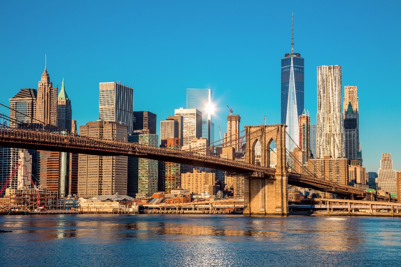 Manhattan skyline with golden sunlight. The Brooklyn Bridge is in the midground, and the blue water is in the foreground.