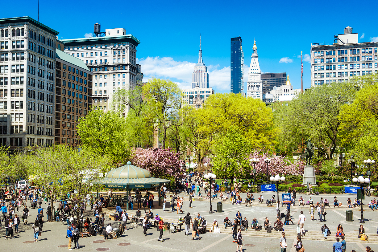 A NYC plaza filled with people milling about in the foreground with trees and buildings in the background