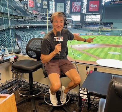 Picture of a man sitting on a stool, holding a microphone in the Chase Field announcer’s booth that overlooks the baseball diamond field below.