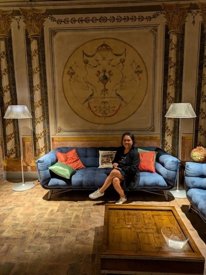 Woman relaxing on a blue sofa with green and orange pillows, two floor lamps and stone flooring with a coffee table in the foreground and an ornate tapestry in the background.