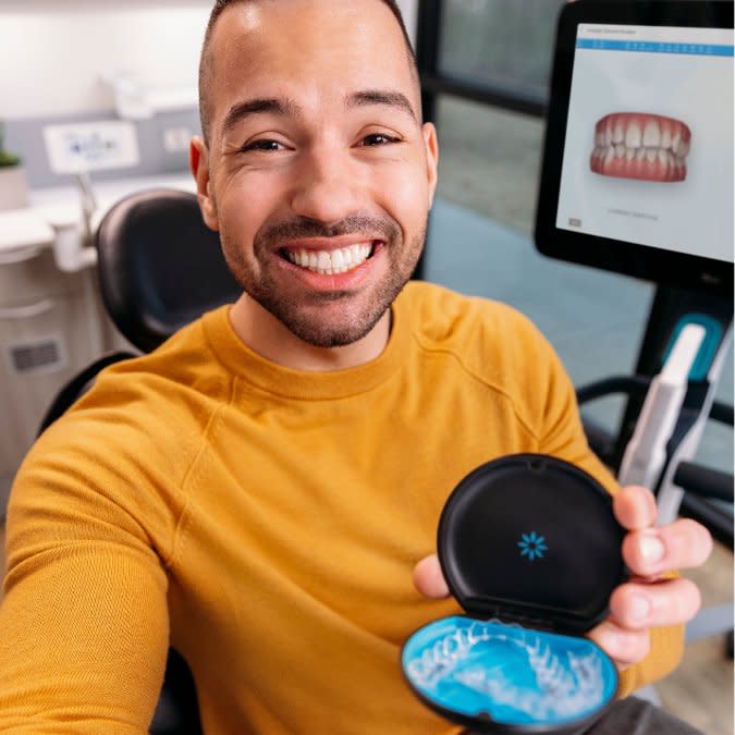 Mutlu bir kadın, arka planda iTero™ tarayıcı varken Invisalign® şeffaf plaklarını tutarak selfie çekiyor