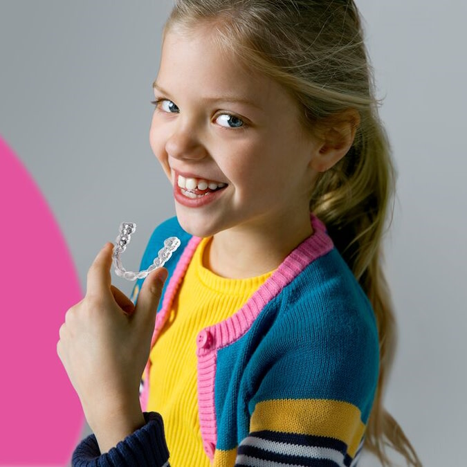 Mała blond dziewczynka uśmiecha się i trzyma przezroczyste nakładki Invisalign®