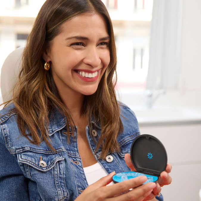 Una ragazza con una giacca di jeans sorride e tiene in mano gli aligner Invisalign®