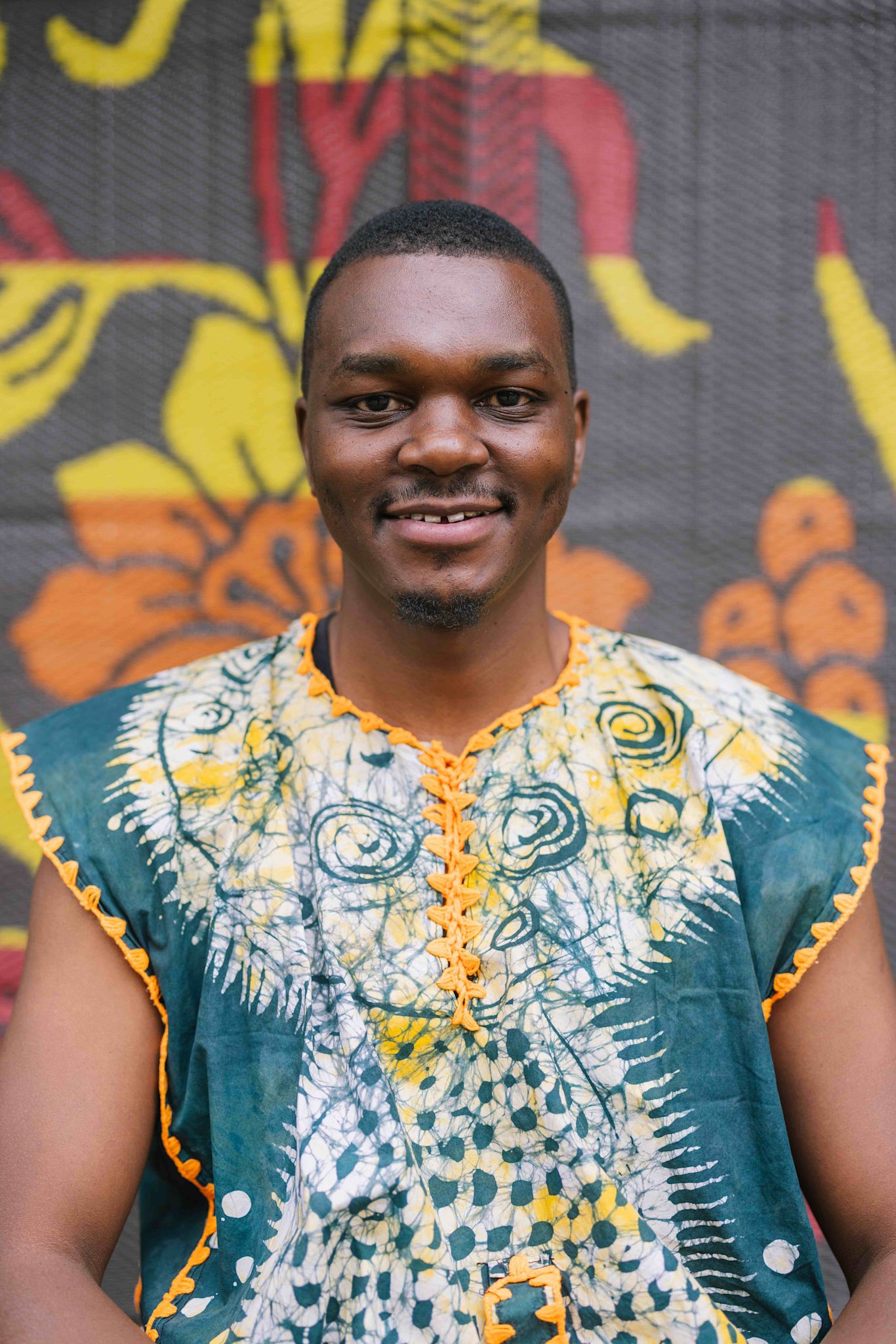 A person smiles at the camera while wearing a green, yellow, and white patterned top with orange trim. They stand in front of a colorful backdrop featuring abstract floral shapes.