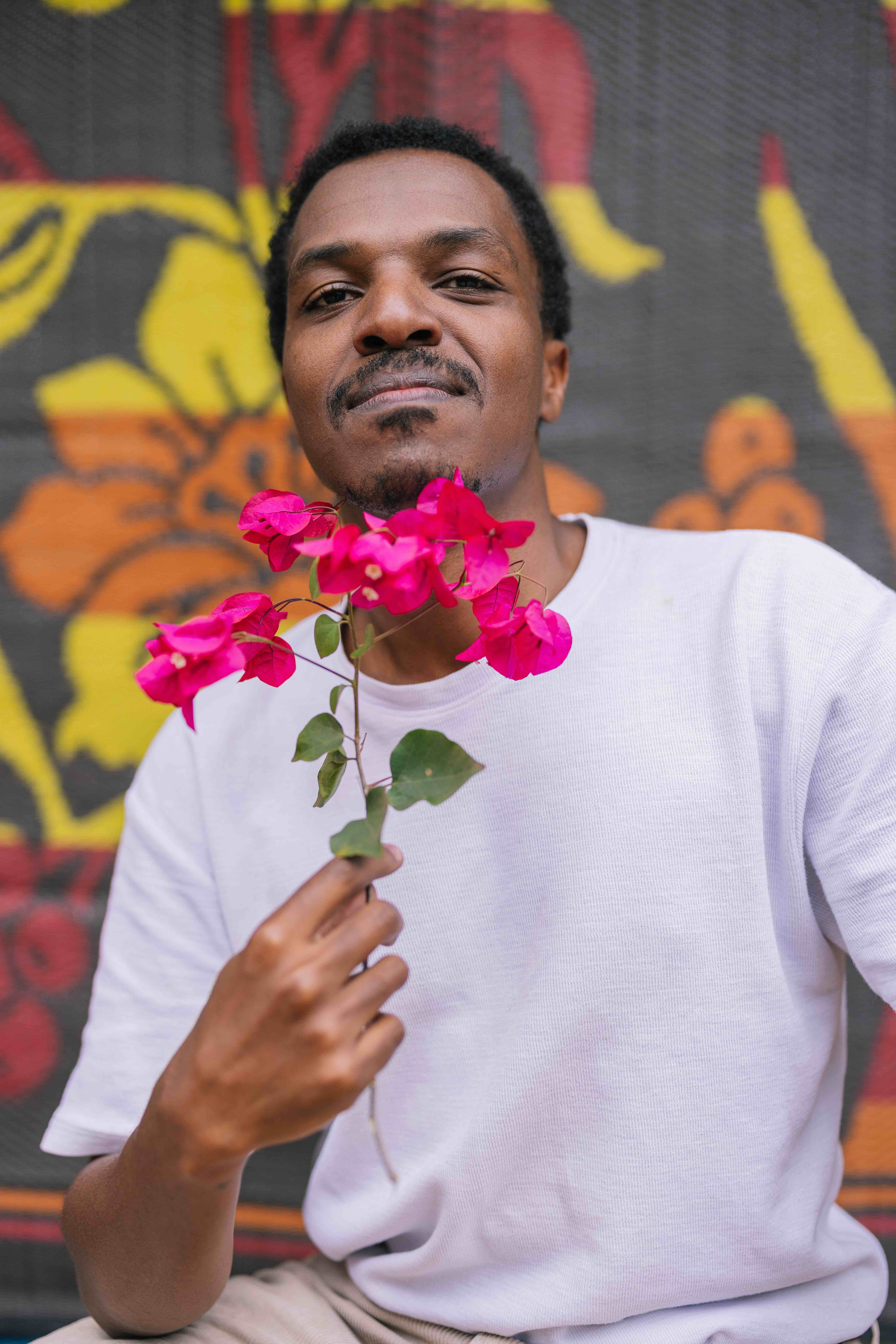 A person in a white shirt holds a small branch of bright pink flowers close to their face, looking calmly at the camera. A colorful patterned backdrop with yellow and orange shapes is visible behind them.