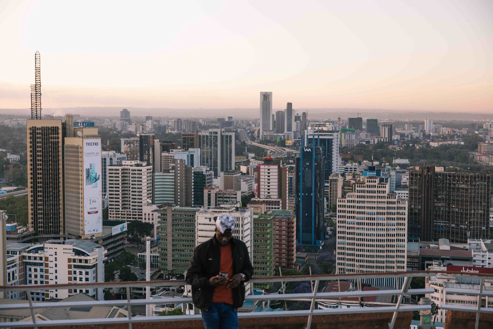 “A wide view of Nairobi’s skyline at dusk, with tall buildings and dense city blocks stretching into the distance. The sky is soft with pastel light. In the foreground, a person stands slightly out of focus on a rooftop, looking down at their phone.