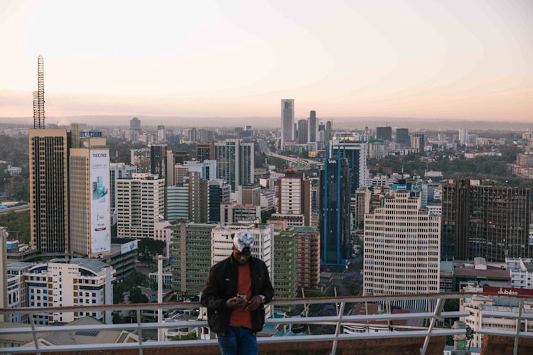 “A wide view of Nairobi’s skyline at dusk, with tall buildings and dense city blocks stretching into the distance. The sky is soft with pastel light. In the foreground, a person stands slightly out of focus on a rooftop, looking down at their phone.