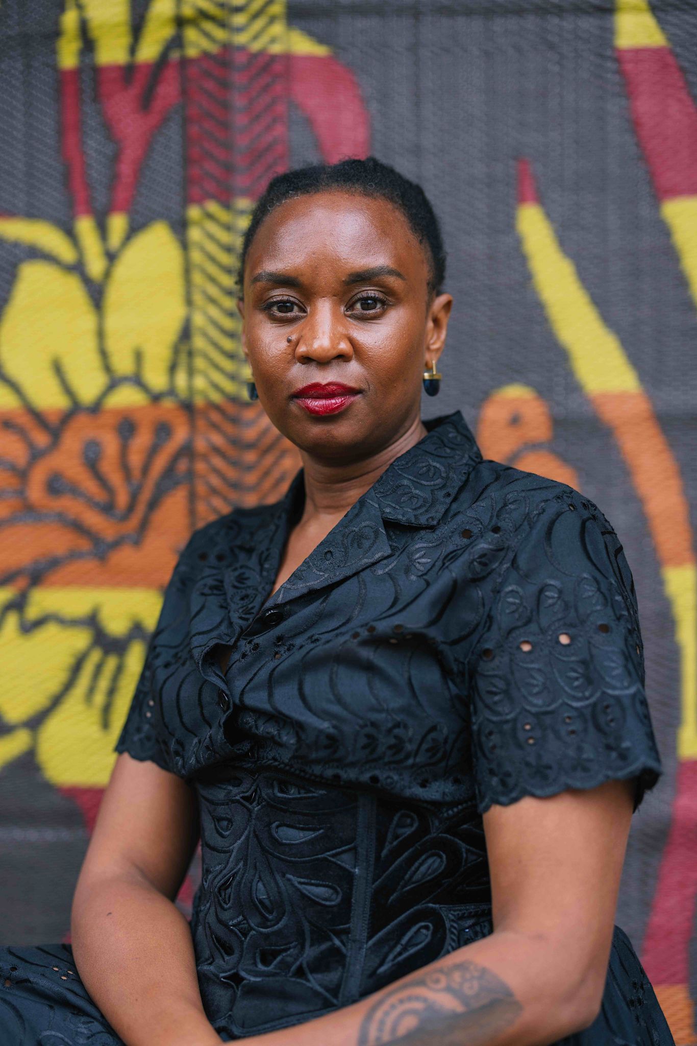 A person sits in front of a colorful patterned backdrop, wearing a black embroidered dress. They look directly at the camera with a steady, composed expression. They wear small earrings and bright red lipstick. A tattoo with circular motifs is visible on their forearm.