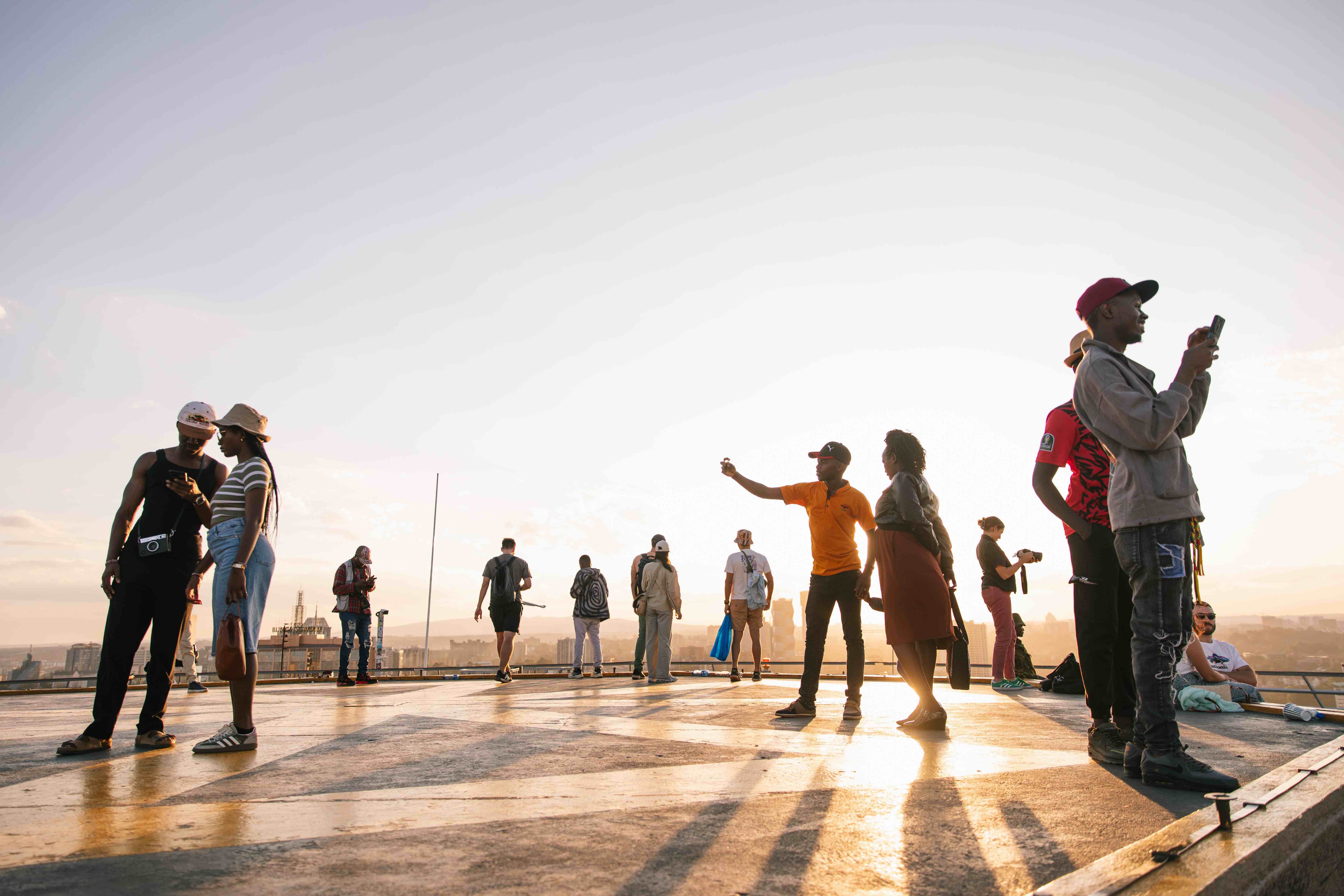 “A group of people stand and walk across a rooftop at sunset, some taking photos and others talking or looking out over the city. Long shadows stretch across the ground as warm golden light fills the scene.
