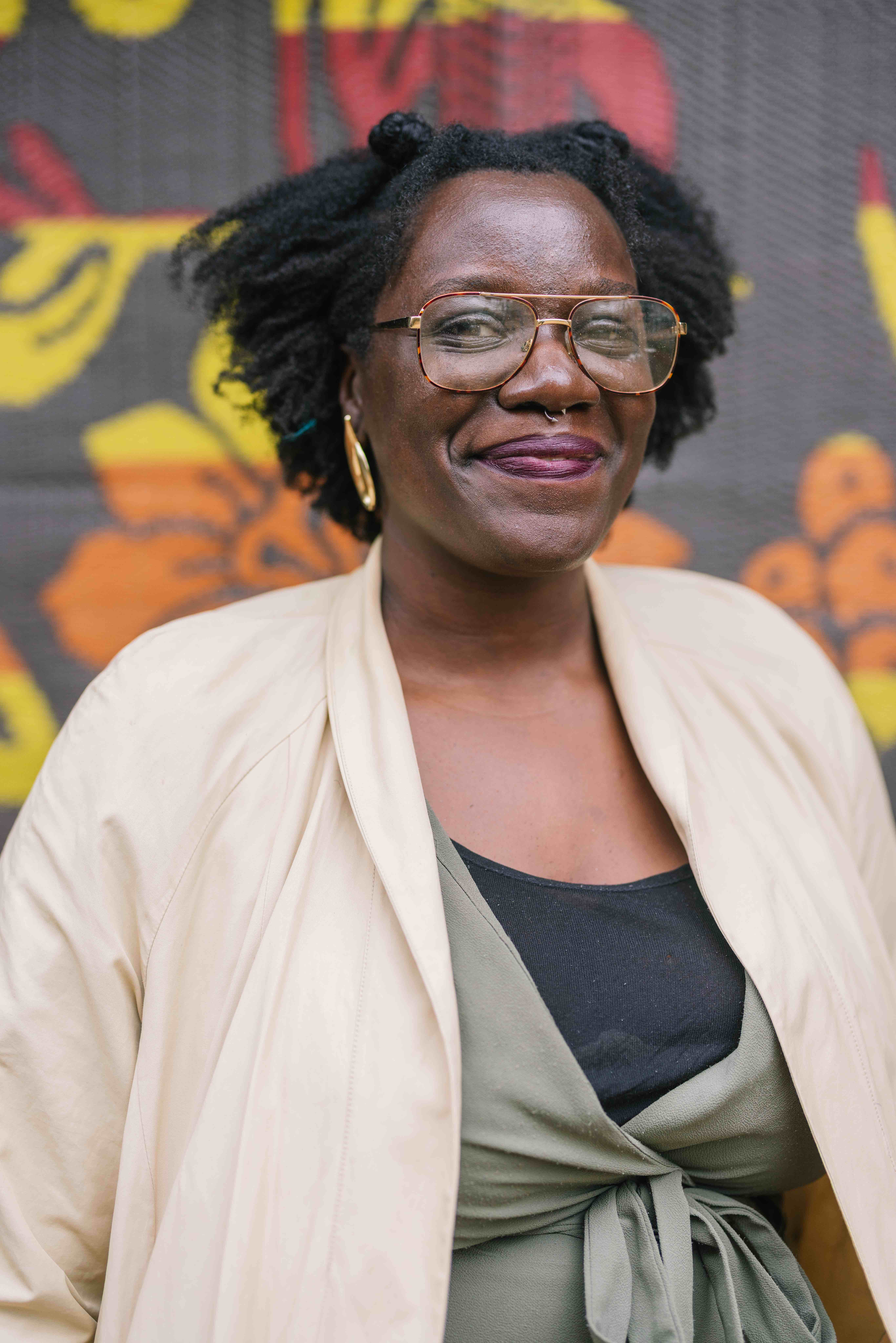 A person smiles warmly at the camera, wearing large glasses, gold hoop earrings, and a cream-colored jacket over a green wrap top. A colorful patterned backdrop with bold yellow and orange shapes is behind them.