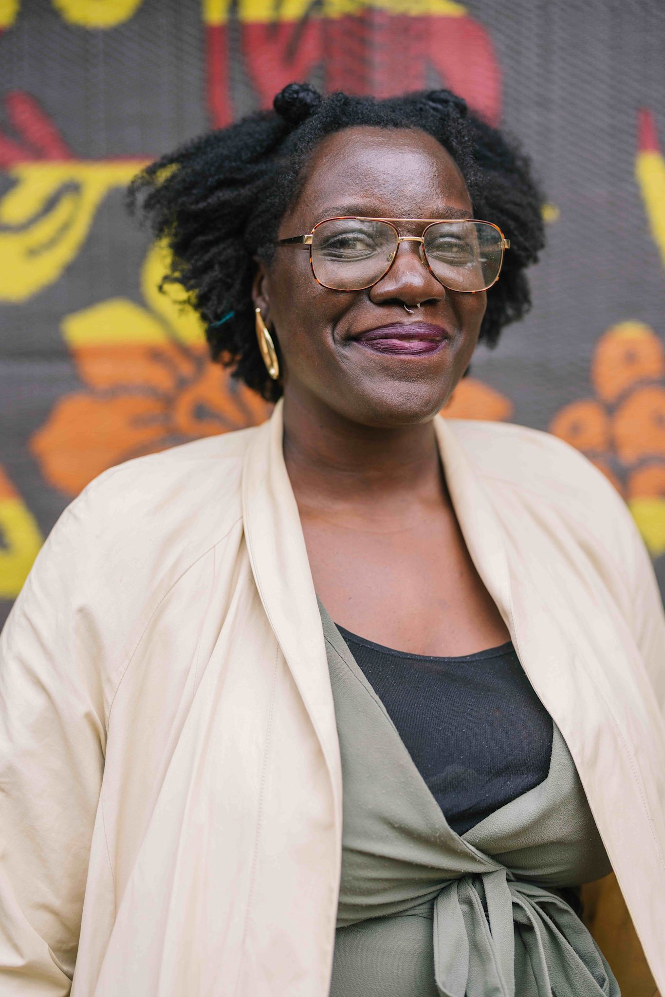 A person smiles warmly at the camera, wearing large glasses, gold hoop earrings, and a cream-colored jacket over a green wrap top. A colorful patterned backdrop with bold yellow and orange shapes is behind them.