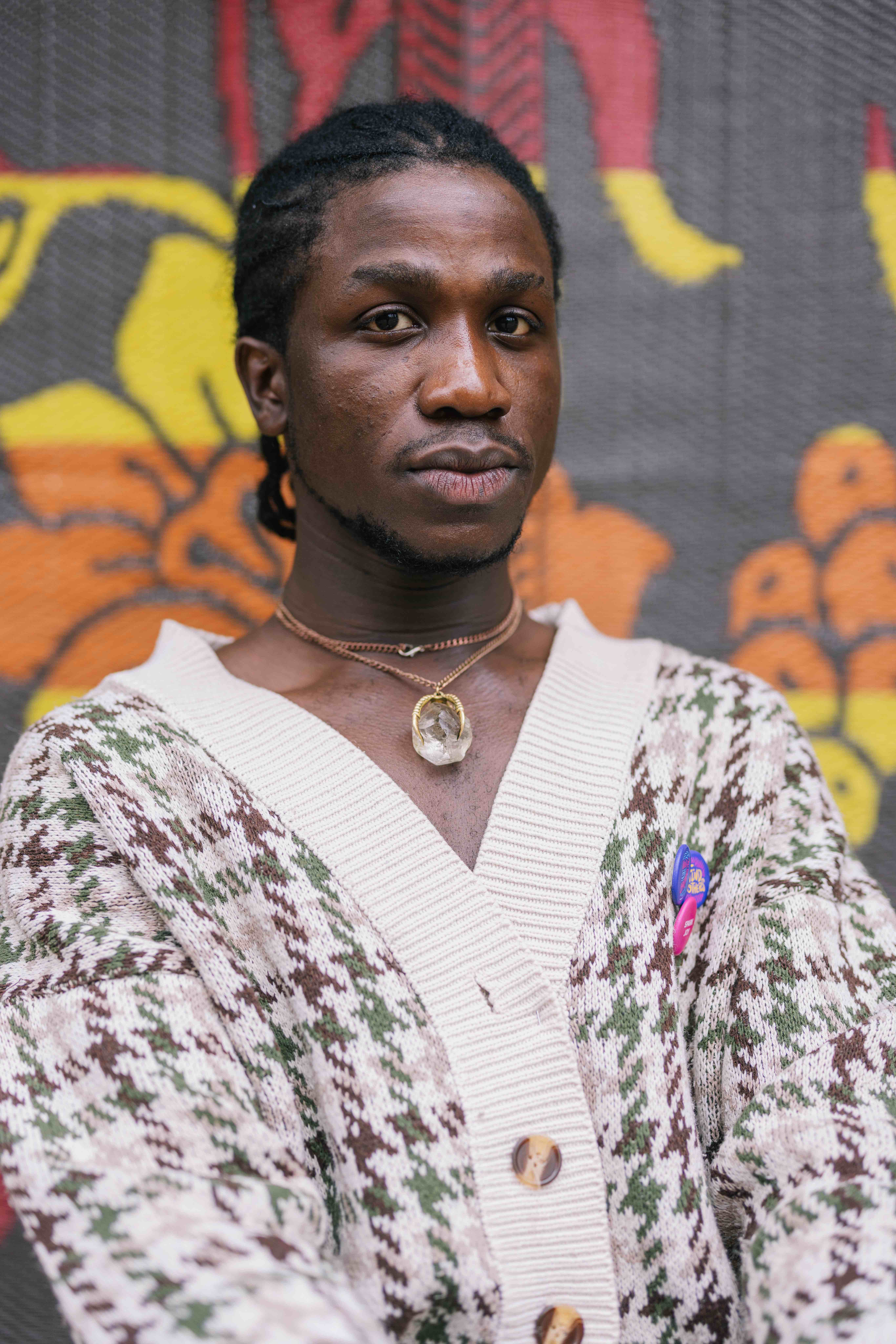 A person looks directly at the camera with a calm, serious expression. They wear a cream and green patterned cardigan, layered necklaces including one with a large crystal pendant, and small colorful pins. A vibrant patterned backdrop with yellow and orange shapes is behind them.