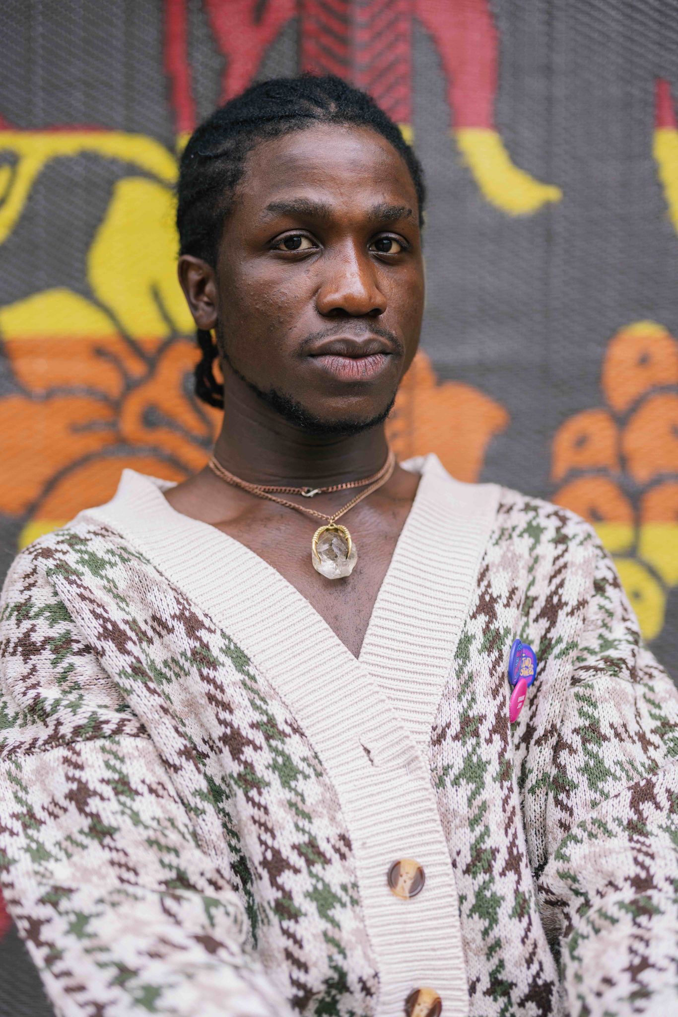 A person looks directly at the camera with a calm, serious expression. They wear a cream and green patterned cardigan, layered necklaces including one with a large crystal pendant, and small colorful pins. A vibrant patterned backdrop with yellow and orange shapes is behind them.