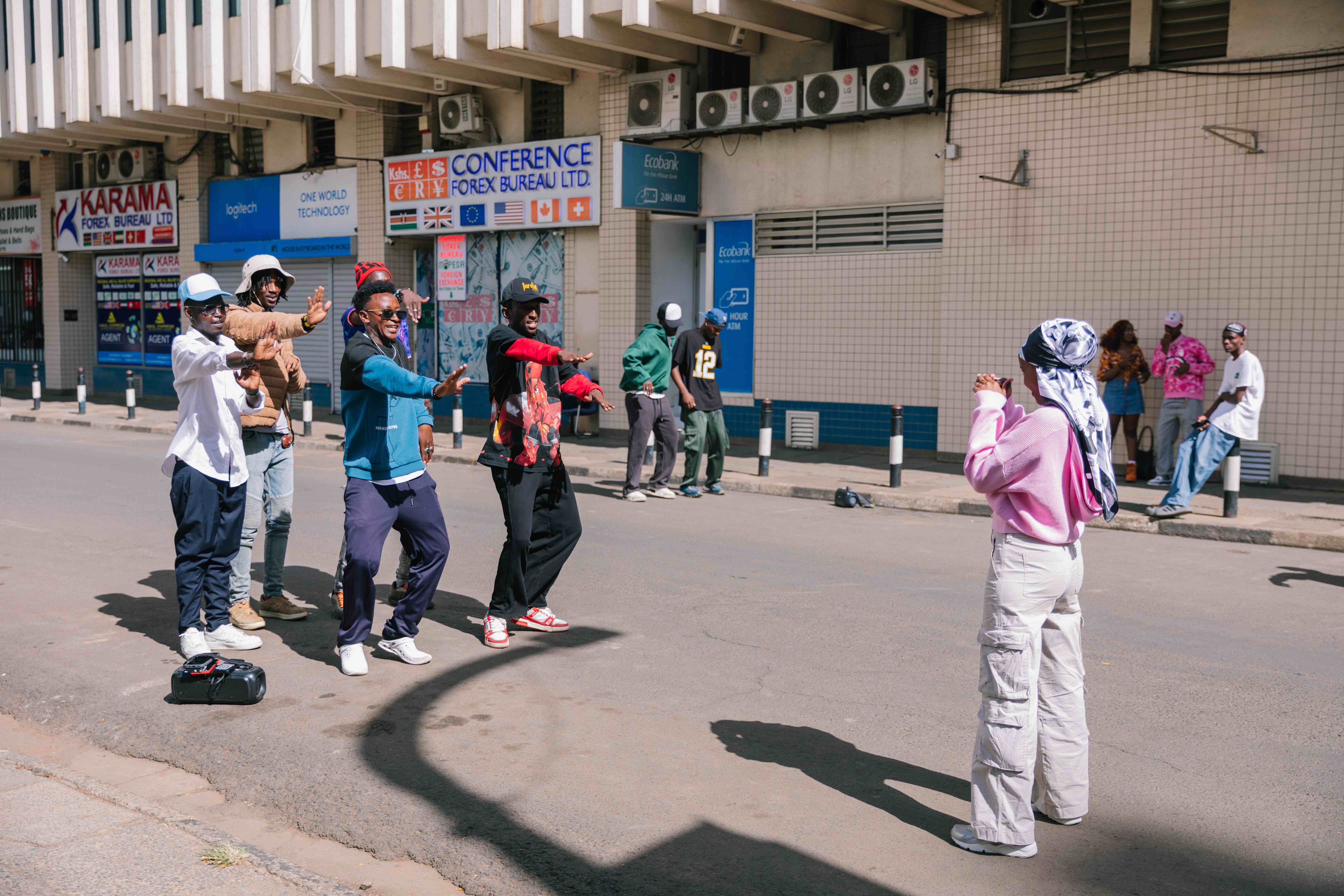 “A group of young people dance in the middle of a city street while another person in a pink sweater and patterned headscarf films them. Others stand along the sidewalk watching. The scene unfolds in bright daylight against storefronts and tiled building façades.