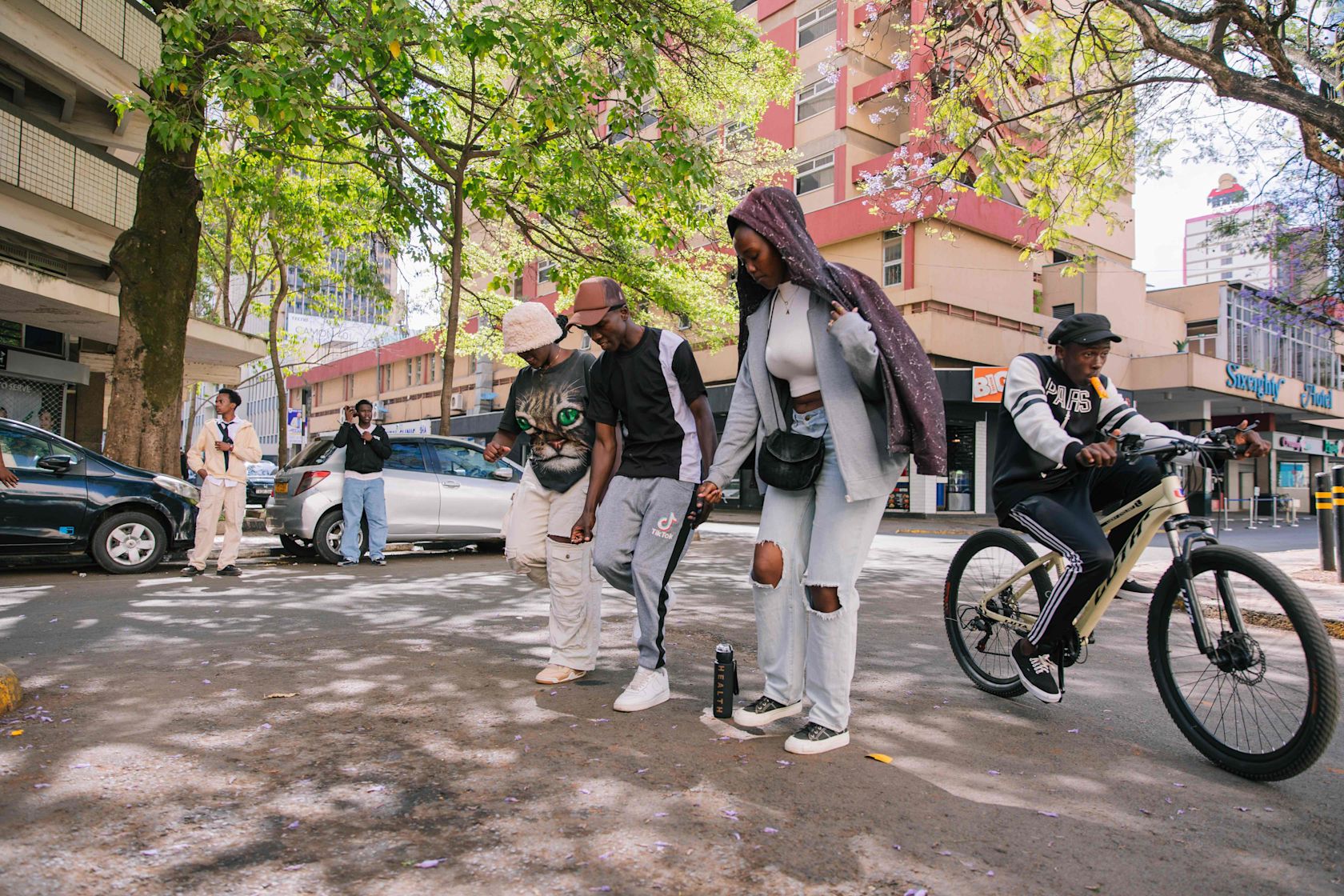 A group of young people walk together down a shaded city street, holding hands and moving in step. One person rides a bicycle beside them. Cars are parked along the road, and tall buildings and trees frame the scene.