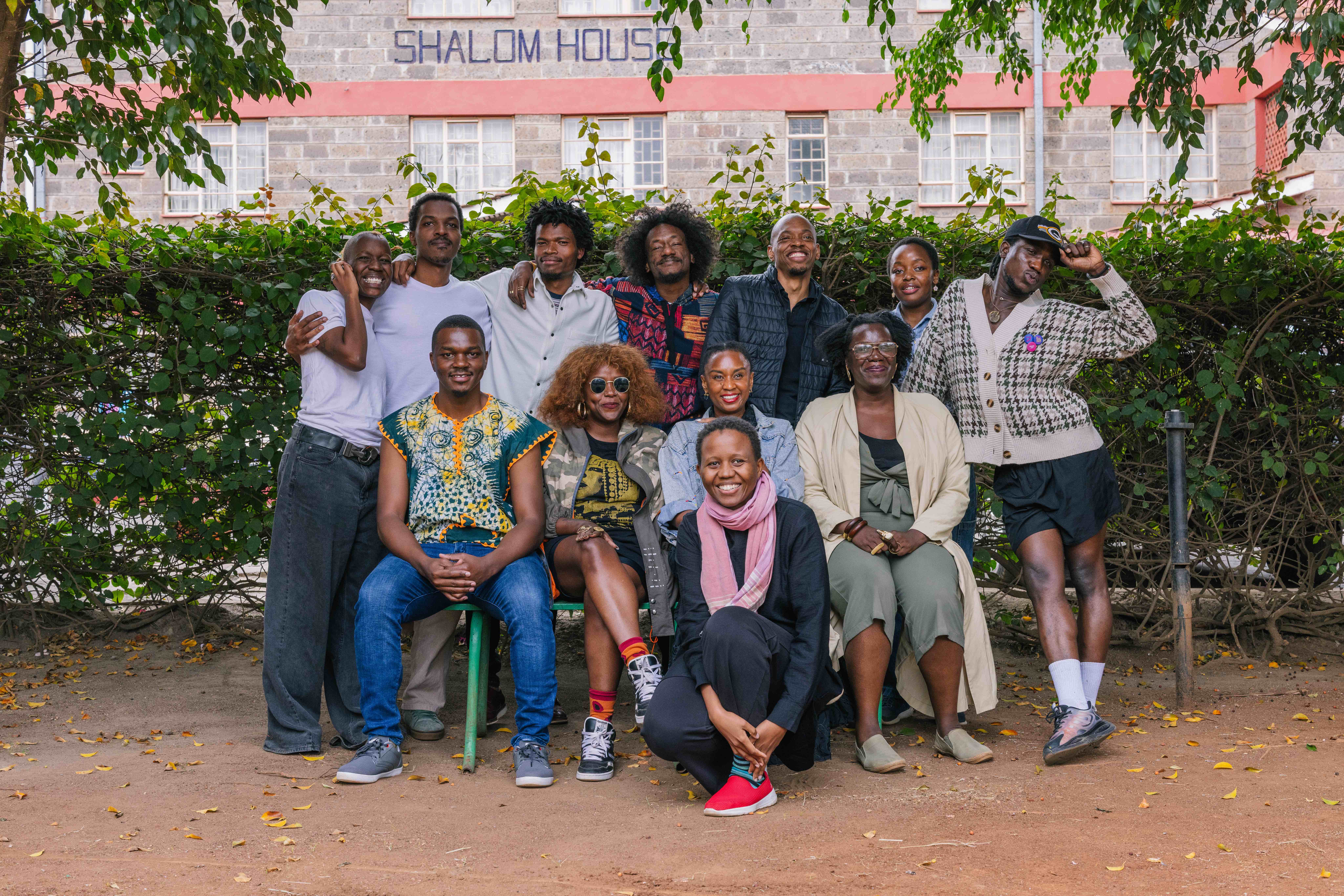 A group of twelve people pose together outdoors in front of a leafy hedge, with a building labeled ‘Shalom House’ in the background. Some are sitting on a bench or stool while others stand behind them, smiling and leaning close. The group appears relaxed and joyful, framed by trees and scattered fallen leaves on the ground.