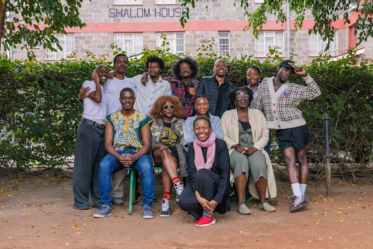 A group of twelve people pose together outdoors in front of a leafy hedge, with a building labeled ‘Shalom House’ in the background. Some are sitting on a bench or stool while others stand behind them, smiling and leaning close. The group appears relaxed and joyful, framed by trees and scattered fallen leaves on the ground.