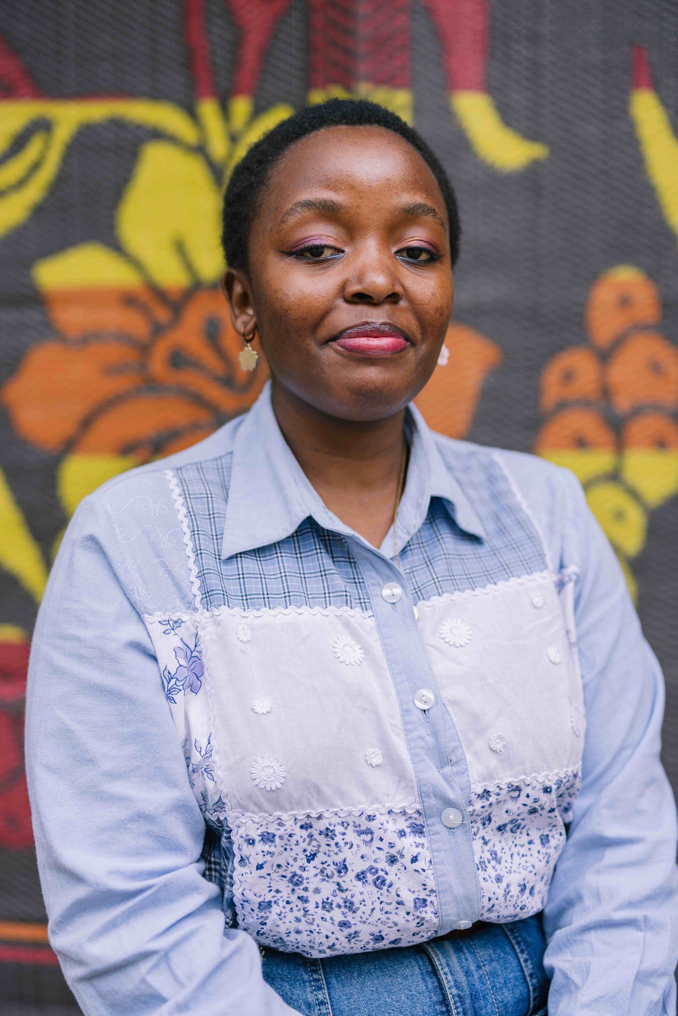 A person with short hair looks softly at the camera, wearing a light blue shirt with white embroidered panels and floral patterns. They have pink eyeshadow, small earrings, and stand in front of a colorful backdrop with bold yellow and orange shapes.