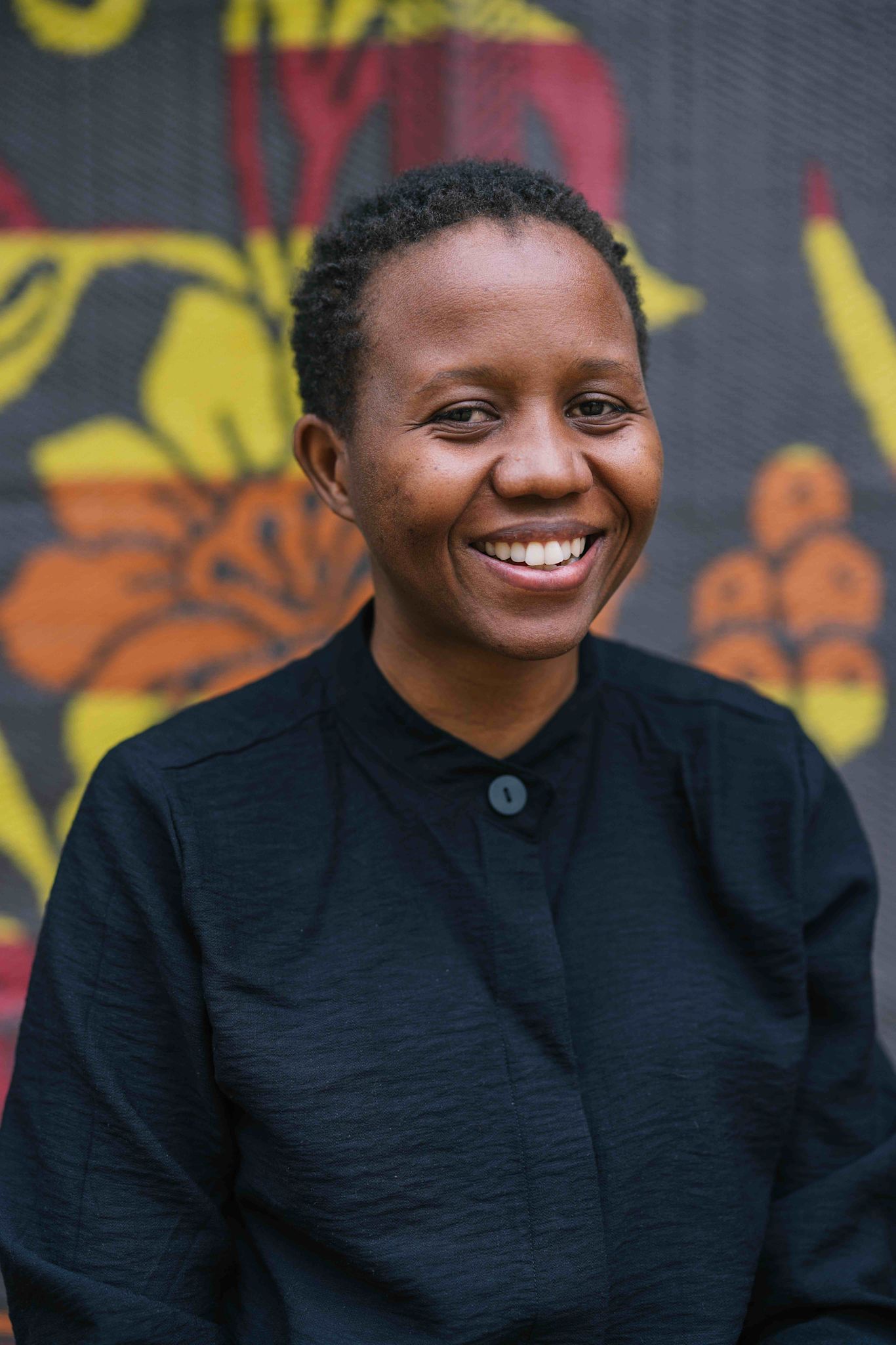 A person smiles brightly at the camera, wearing a black button-up top. They stand in front of a colorful patterned backdrop featuring bold yellow and orange shapes.
