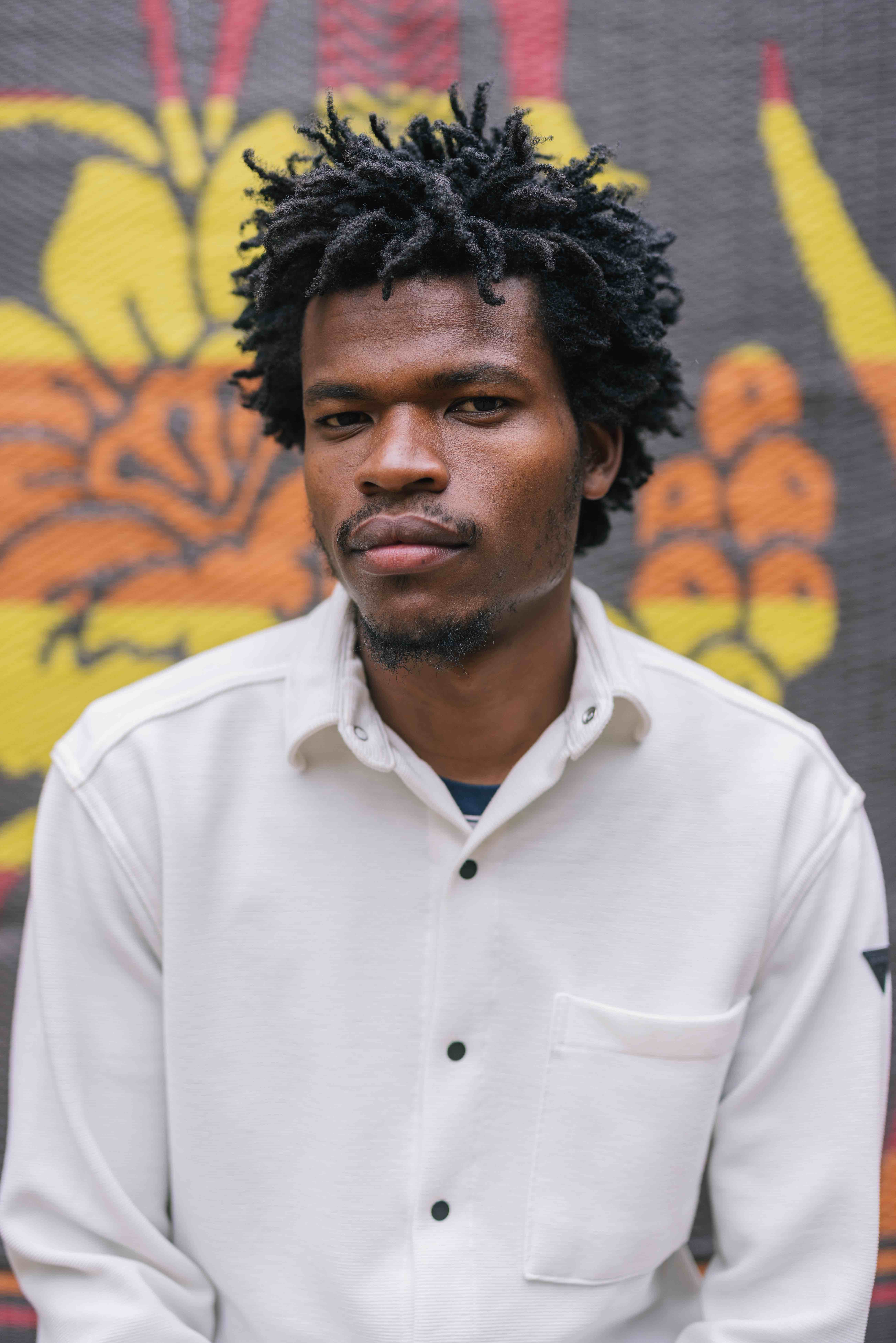 “A person with short, textured hair looks directly at the camera with a neutral expression. They wear a white button-up shirt and stand in front of a colorful patterned backdrop featuring bold yellow, orange, and red shapes.