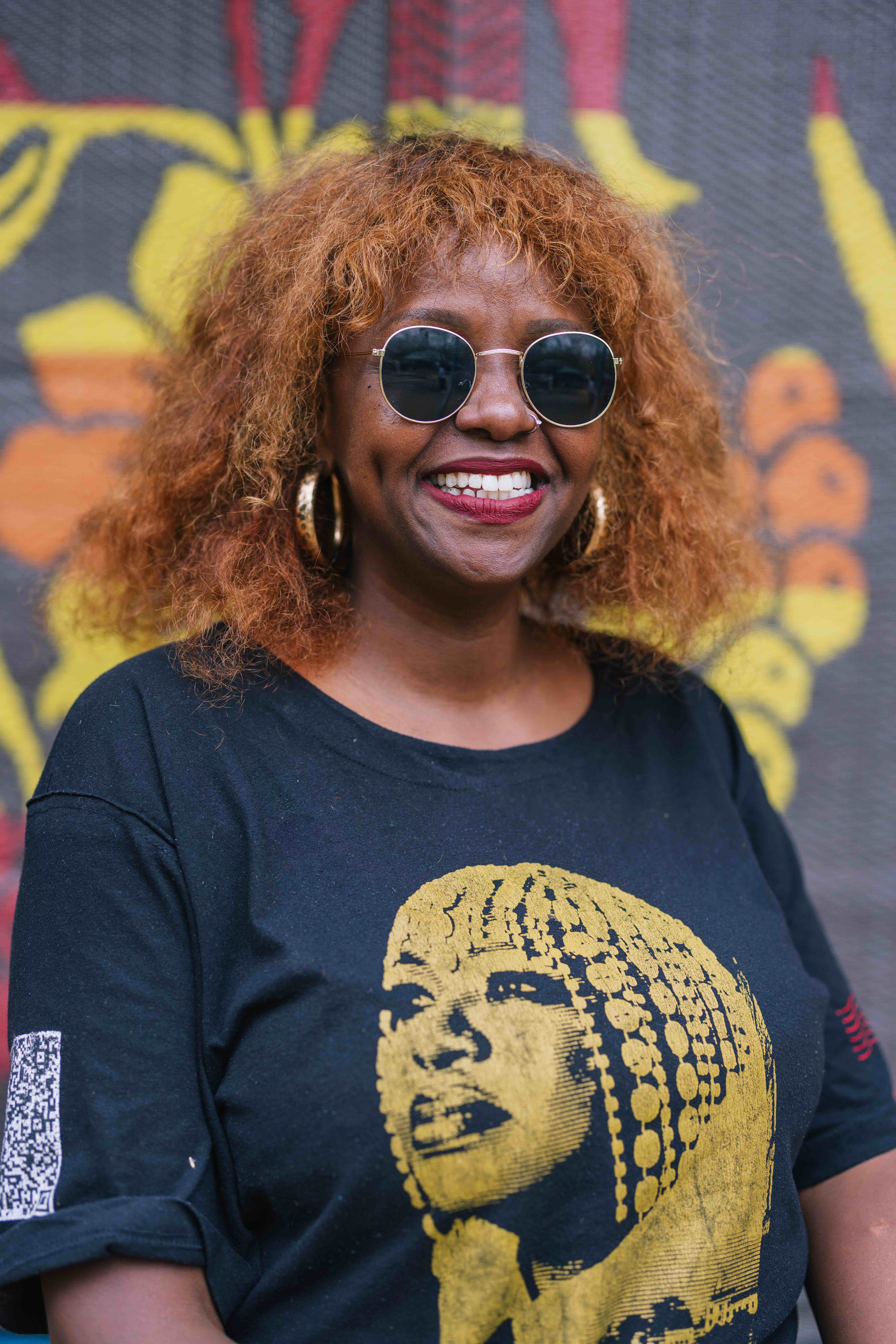 A person smiles at the camera while wearing round sunglasses and large hoop earrings. They are dressed in a black T-shirt featuring a gold graphic portrait. A colorful patterned backdrop is visible behind them.