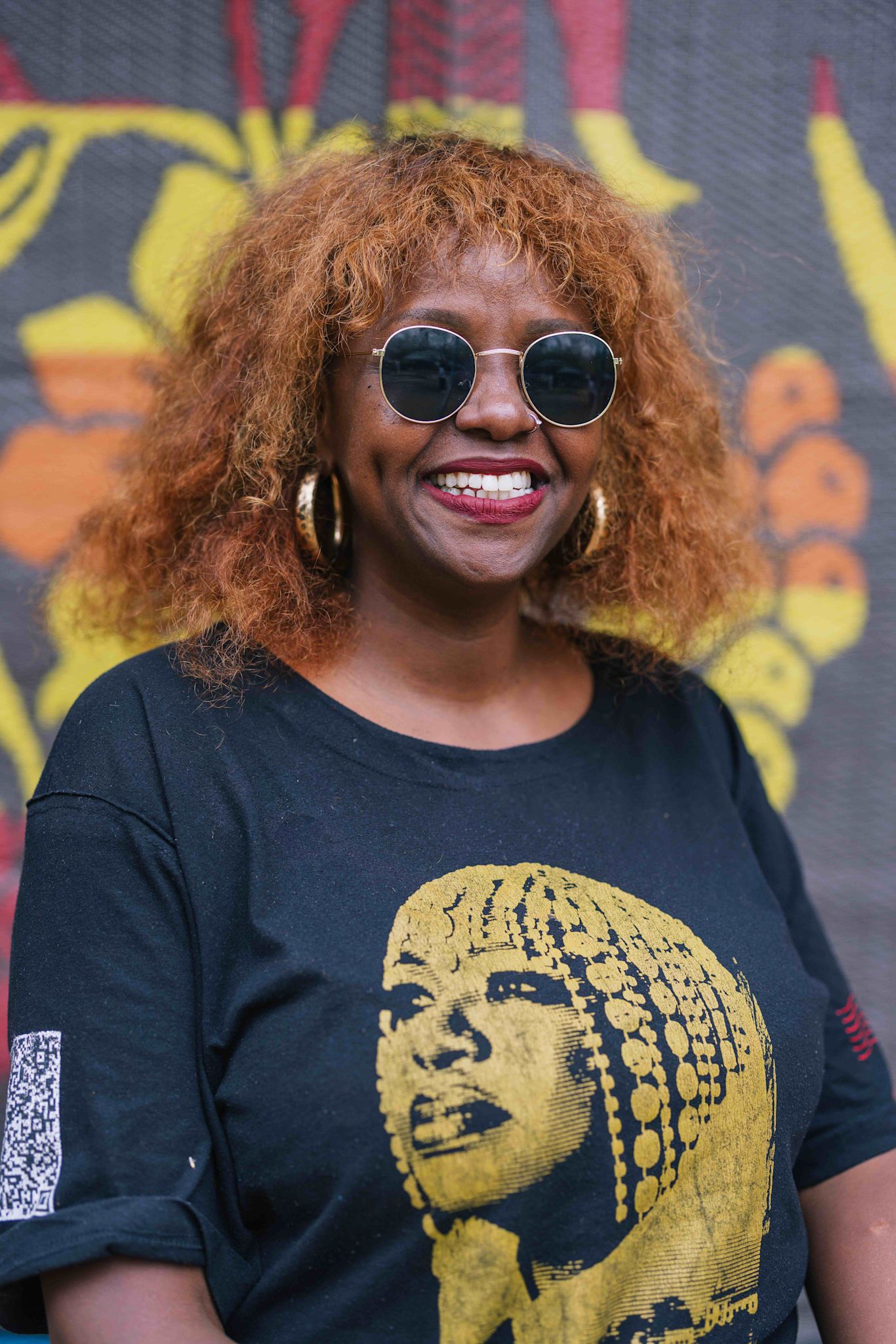A person smiles at the camera while wearing round sunglasses and large hoop earrings. They are dressed in a black T-shirt featuring a gold graphic portrait. A colorful patterned backdrop is visible behind them.