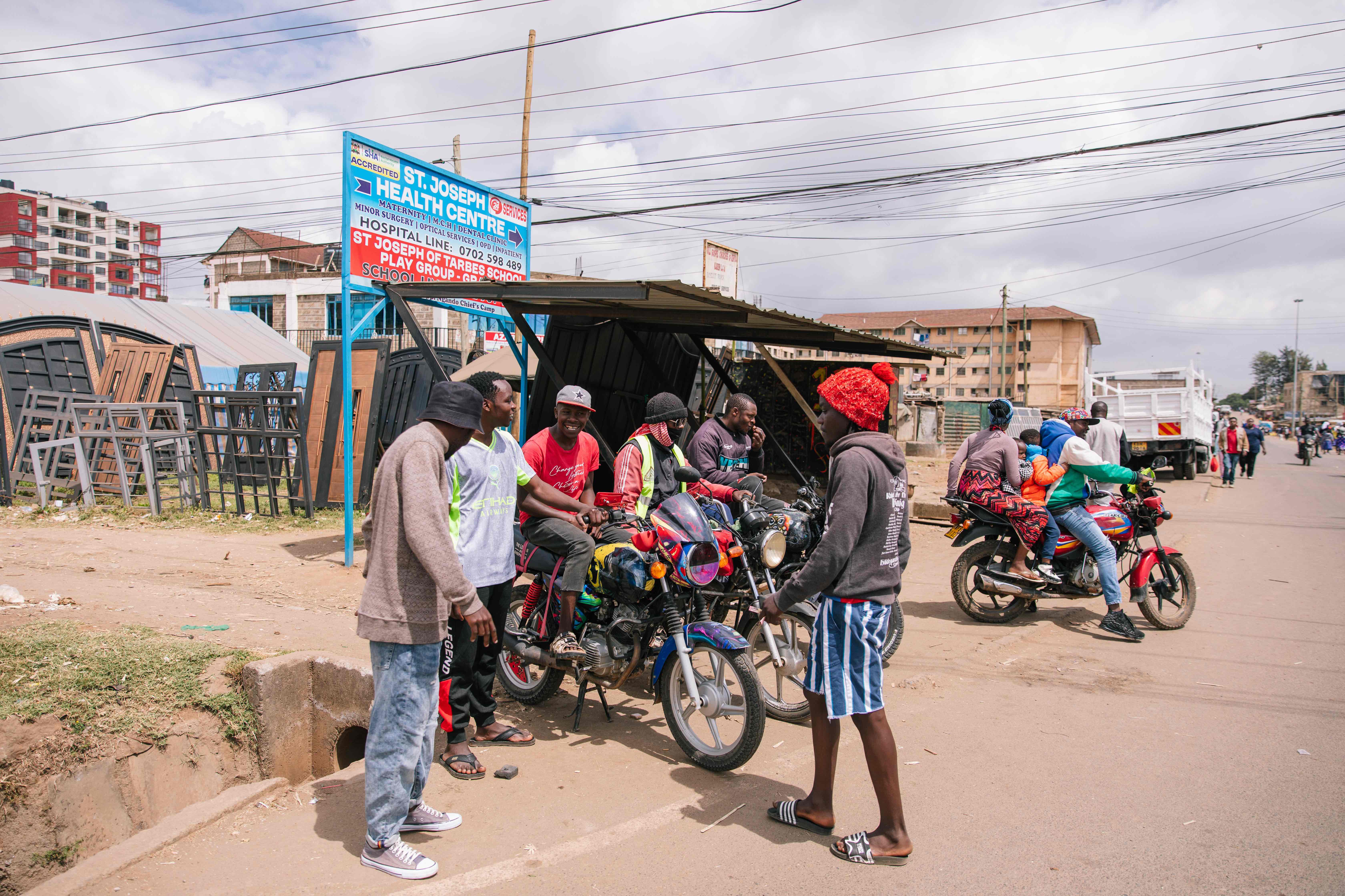 “A group of motorcycle taxi riders gather and talk beside a roadside shelter, with several motorbikes parked in front of them. Nearby, another motorbike carries multiple passengers, including children. The scene takes place along a busy street lined with metal gates, small shops, and residential buildings under an overcast sky.