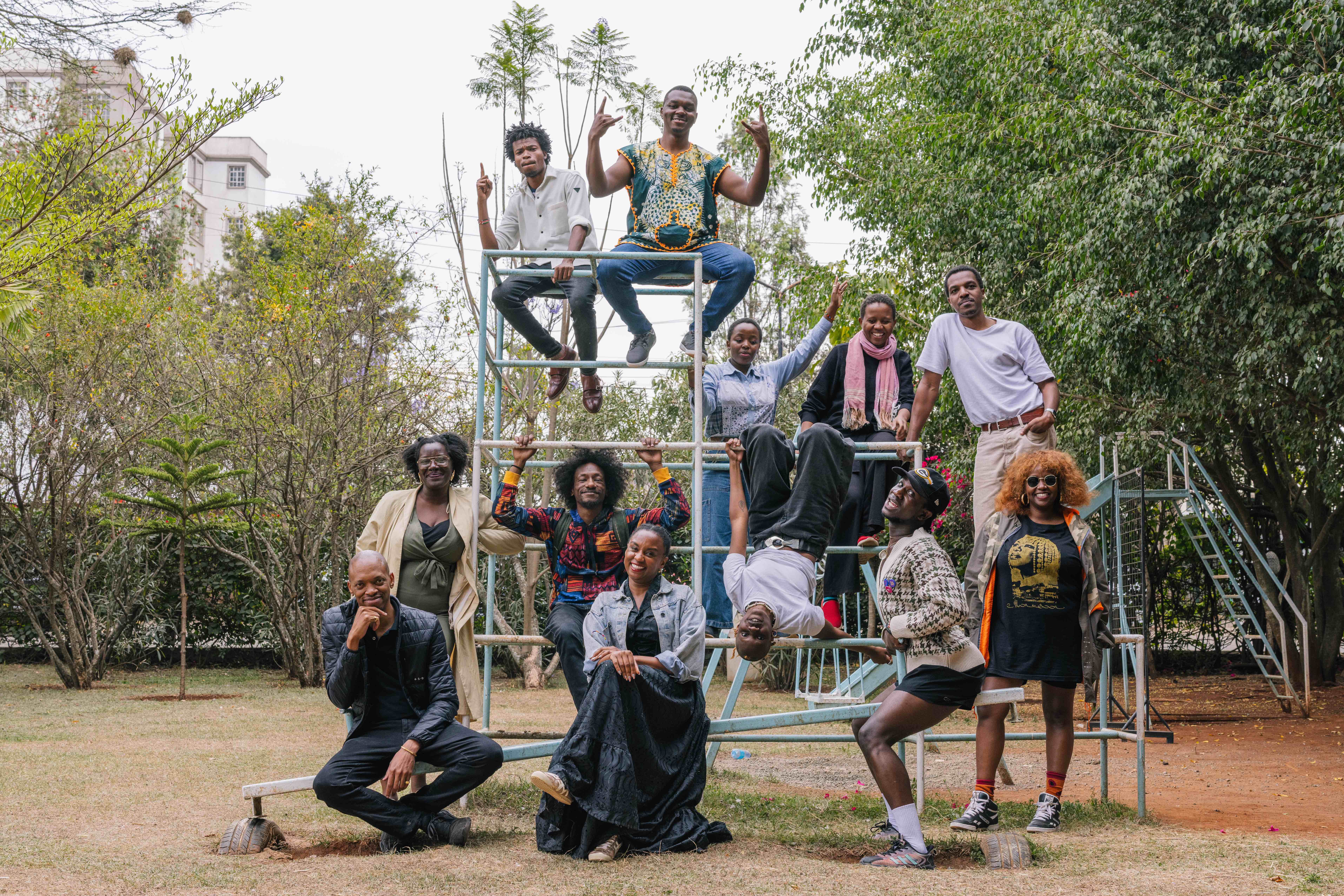 A group of twelve people pose playfully on a metal climbing structure in an outdoor park. Some sit at the top, others stand or lean against the bars, and a few hang upside down or strike expressive poses. They appear joyful and energetic, surrounded by trees and greenery.
