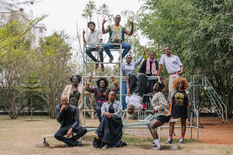 A group of twelve people pose playfully on a metal climbing structure in an outdoor park. Some sit at the top, others stand or lean against the bars, and a few hang upside down or strike expressive poses. They appear joyful and energetic, surrounded by trees and greenery.