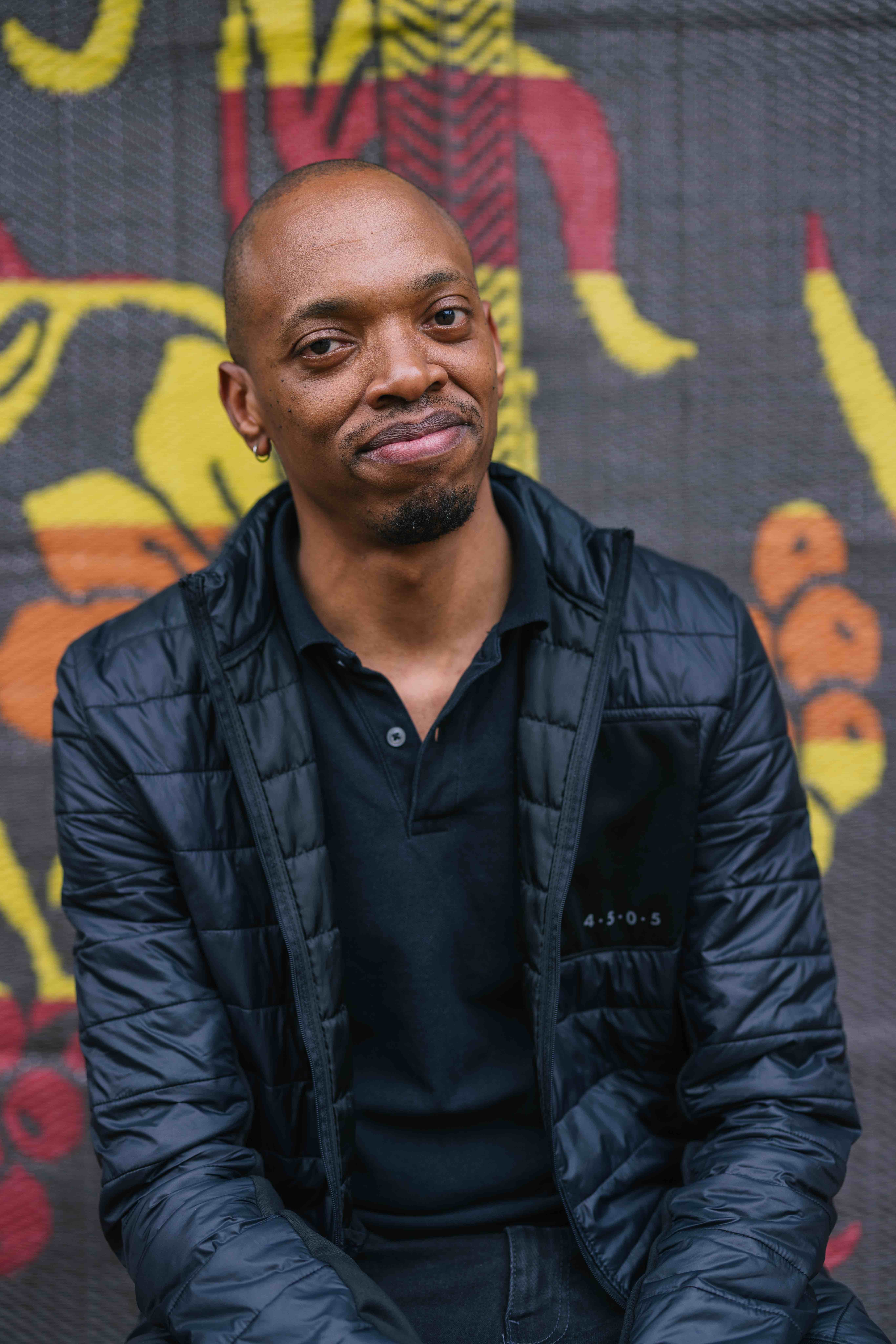 A person wearing a black quilted jacket and black shirt looks toward the camera with a slight smile. They have a shaved head, a small hoop earring, and a short goatee. Behind them is a colorful patterned backdrop with bold yellow, orange, and red shapes.