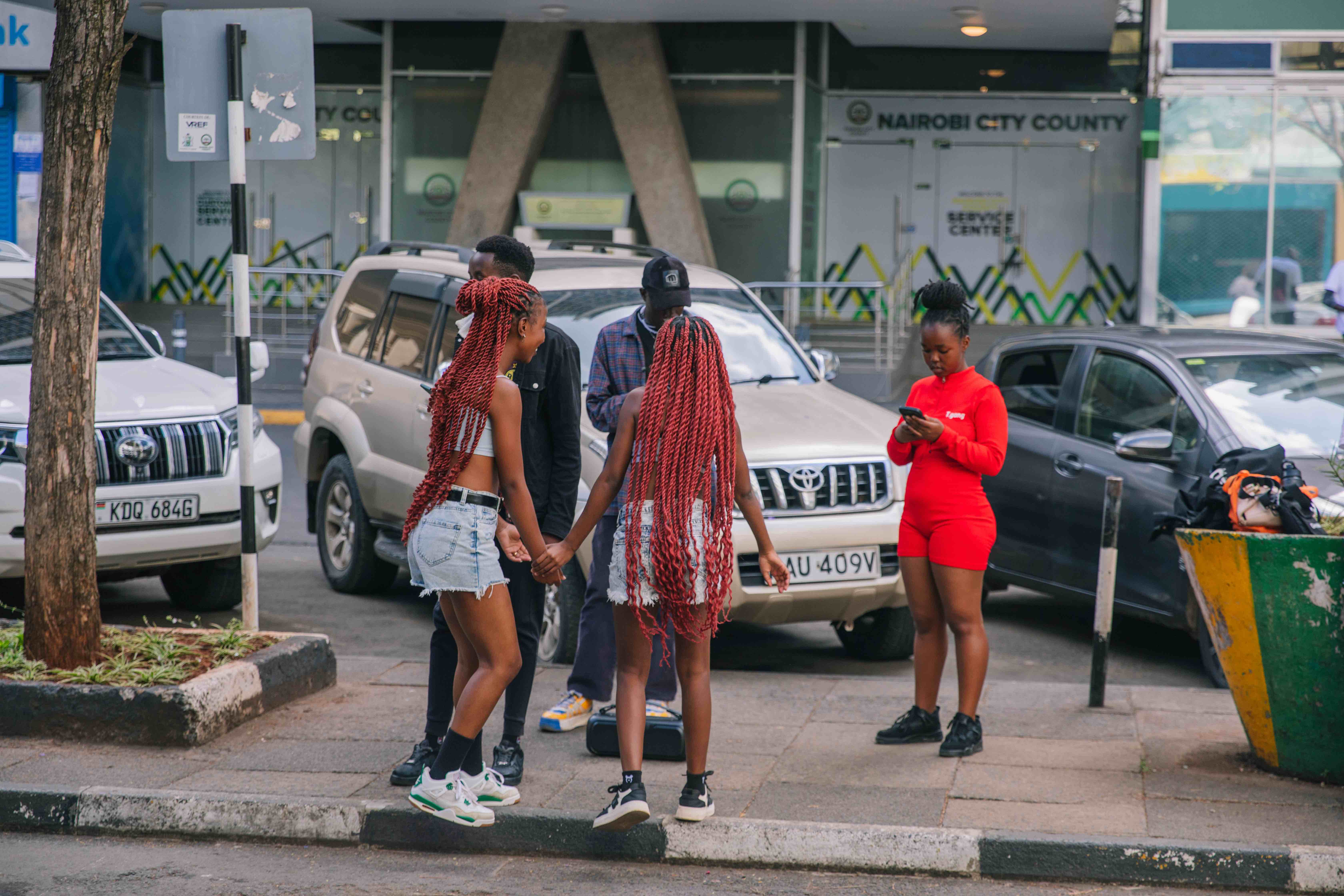 A small group of young people gather on a city sidewalk near parked cars. Two girls with long red braids hold hands and face each other while talking. Nearby, a person in a bright red outfit looks down at their phone. The scene takes place in front of a building marked ‘Nairobi City County.’