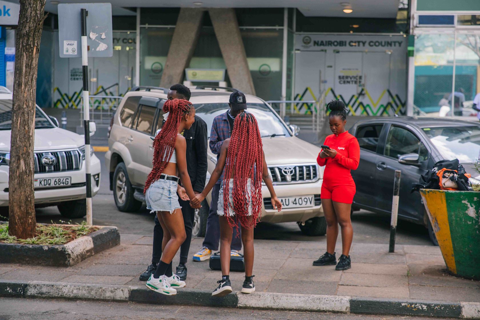 A small group of young people gather on a city sidewalk near parked cars. Two girls with long red braids hold hands and face each other while talking. Nearby, a person in a bright red outfit looks down at their phone. The scene takes place in front of a building marked ‘Nairobi City County.’