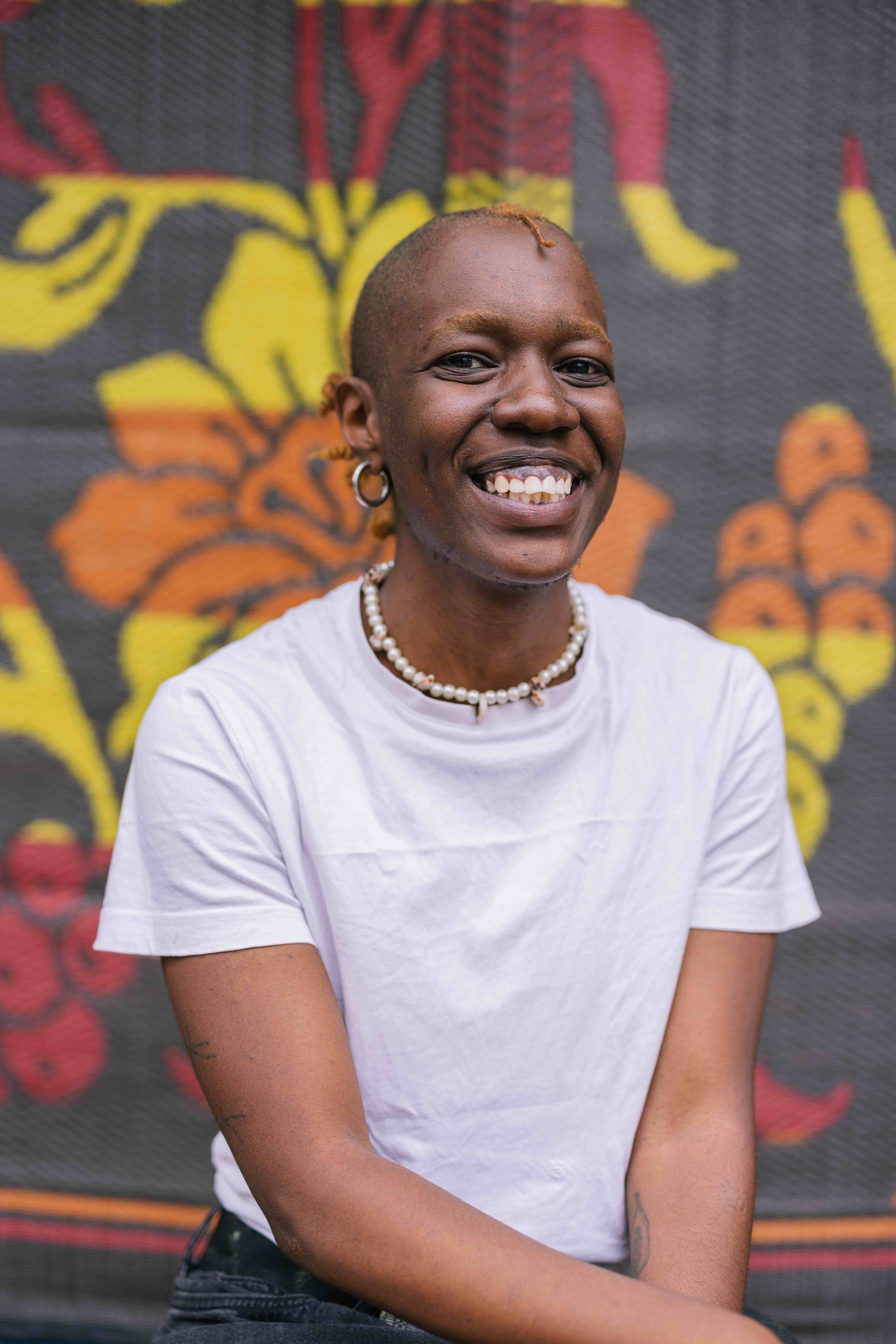 “A person with short, light-colored hair smiles widely at the camera. They wear a white T-shirt, a beaded necklace, and hoop earrings. A colorful patterned backdrop with bold yellow, orange, and red shapes is behind them.