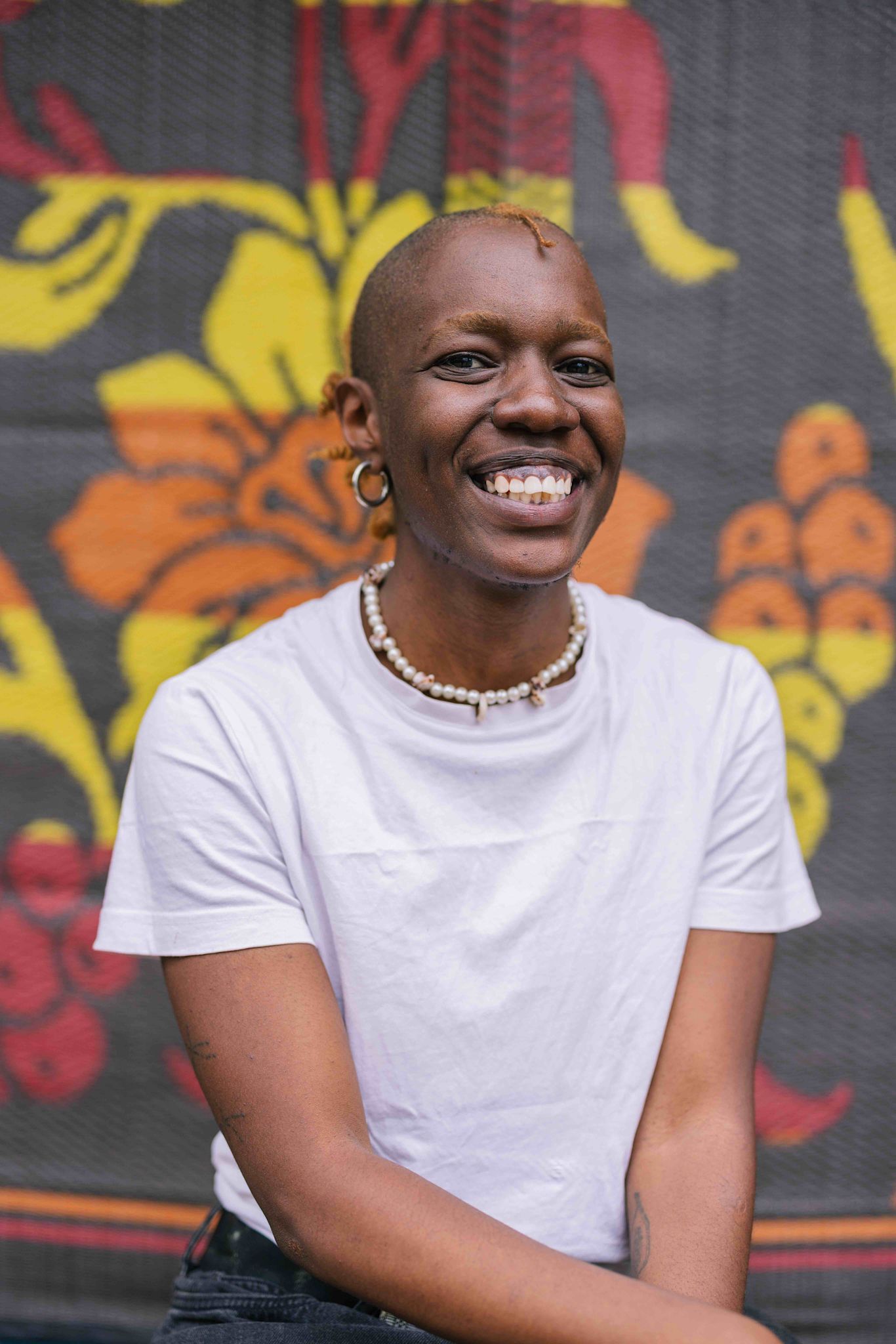 “A person with short, light-colored hair smiles widely at the camera. They wear a white T-shirt, a beaded necklace, and hoop earrings. A colorful patterned backdrop with bold yellow, orange, and red shapes is behind them.