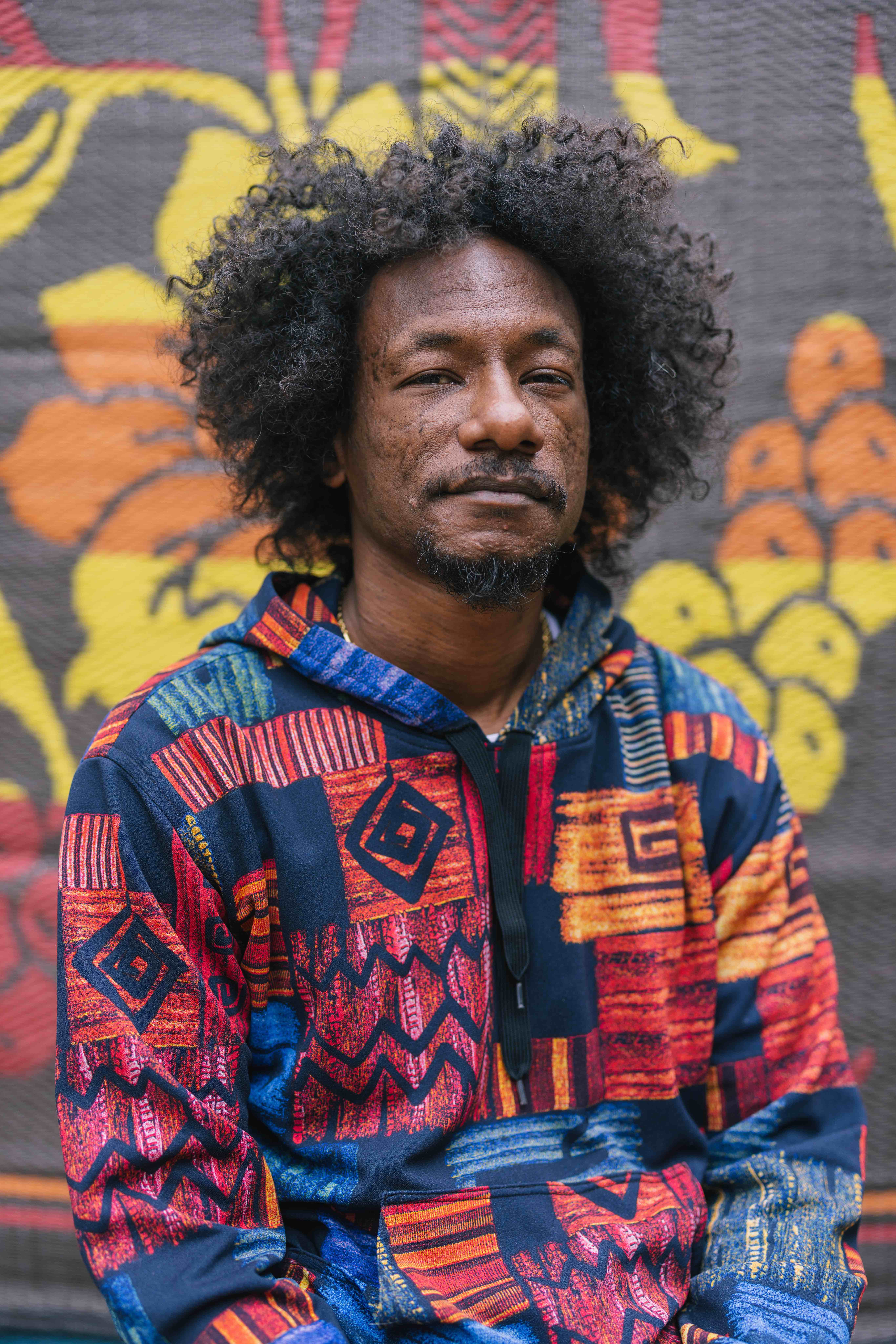 A person sits in front of a colorful patterned backdrop, wearing a vibrant hoodie with red, blue, yellow, and orange geometric designs. They face the camera with a calm, neutral expression.