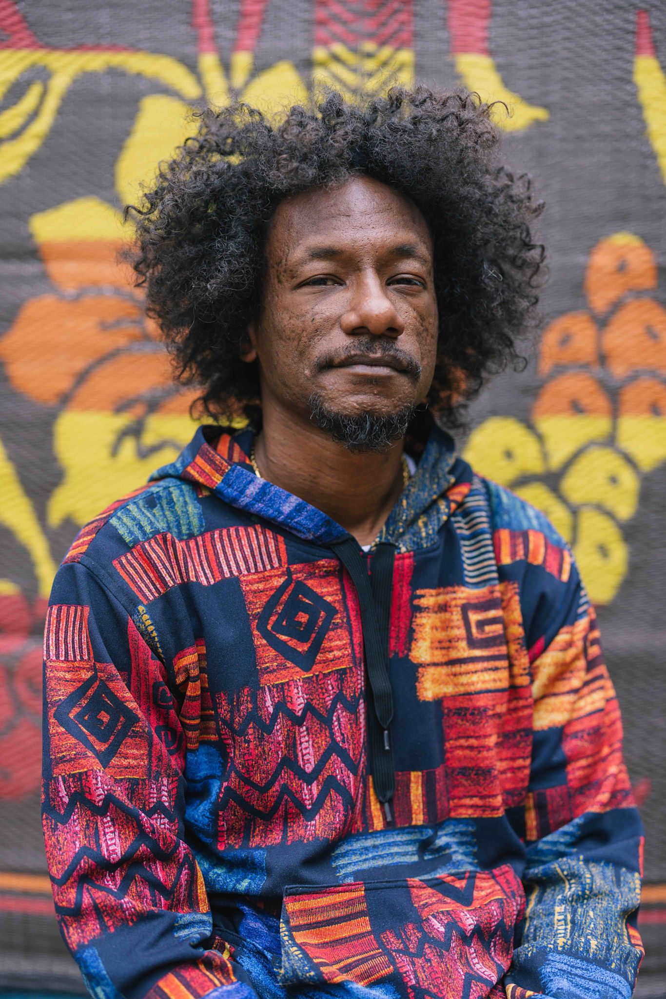 A person sits in front of a colorful patterned backdrop, wearing a vibrant hoodie with red, blue, yellow, and orange geometric designs. They face the camera with a calm, neutral expression.