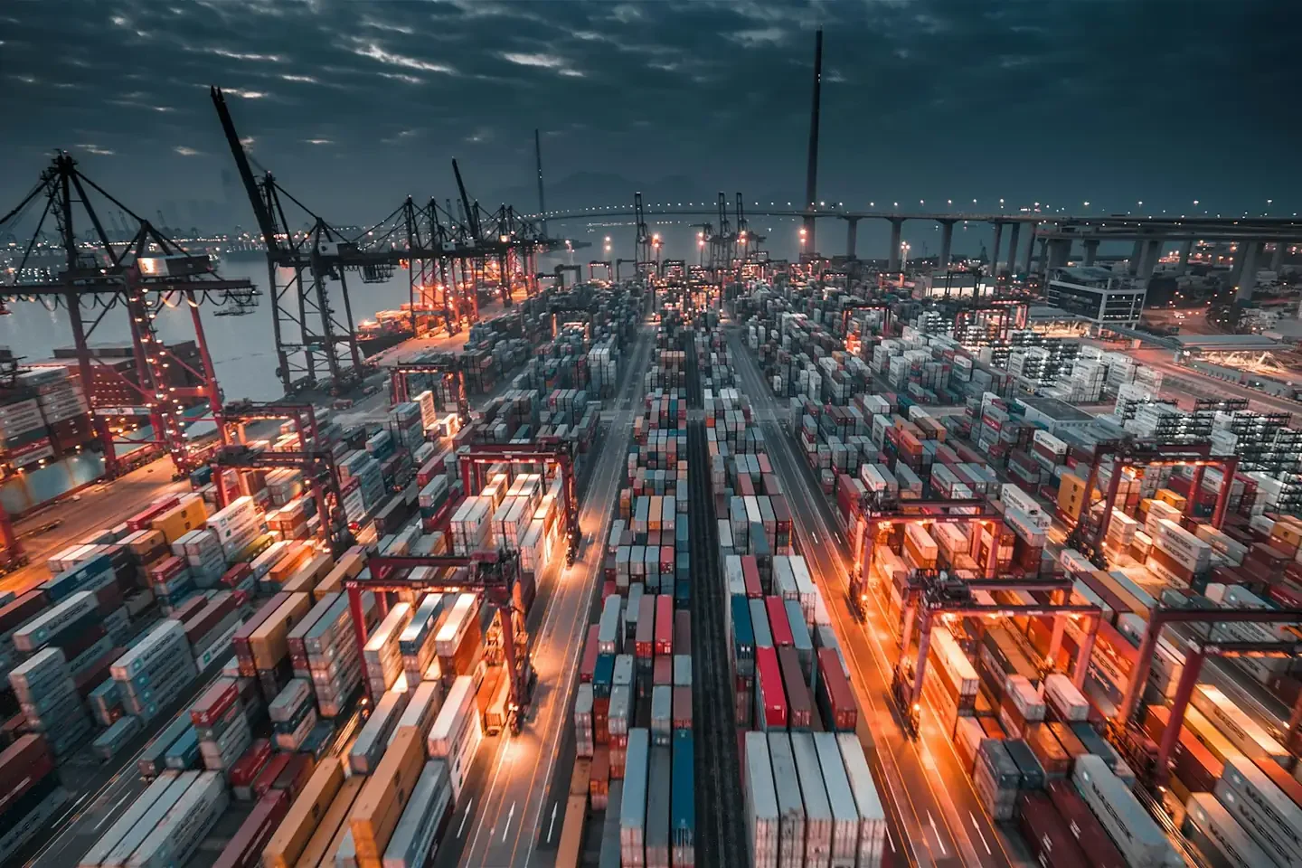 A busy shipping port with stacked containers and cranes under a dark cloudy sky
