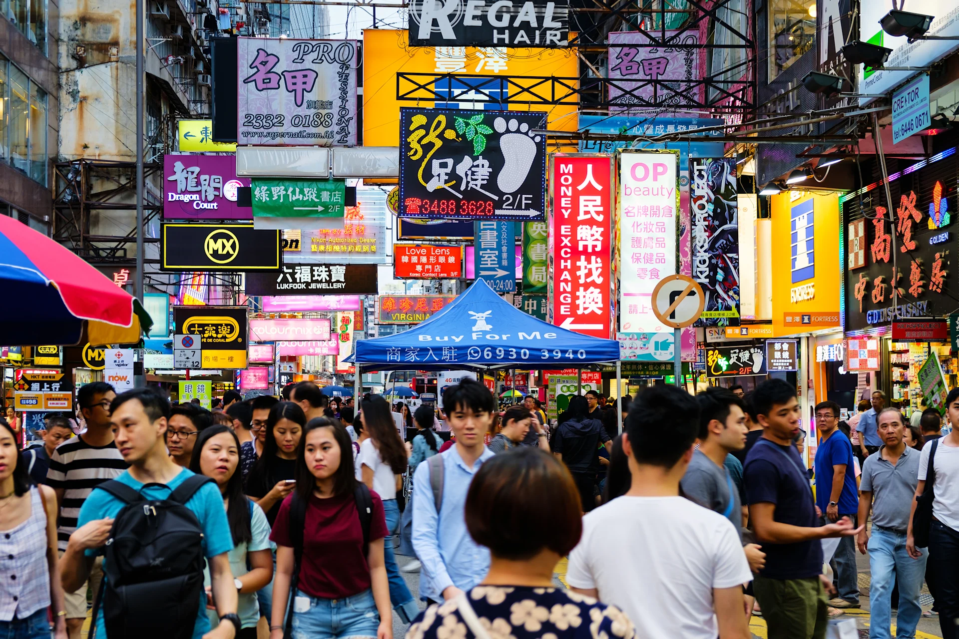 A busy street scene in a vibrant urban area filled with pedestrians. Colourful neon signs and advertisements in various languages illuminate the surroundings