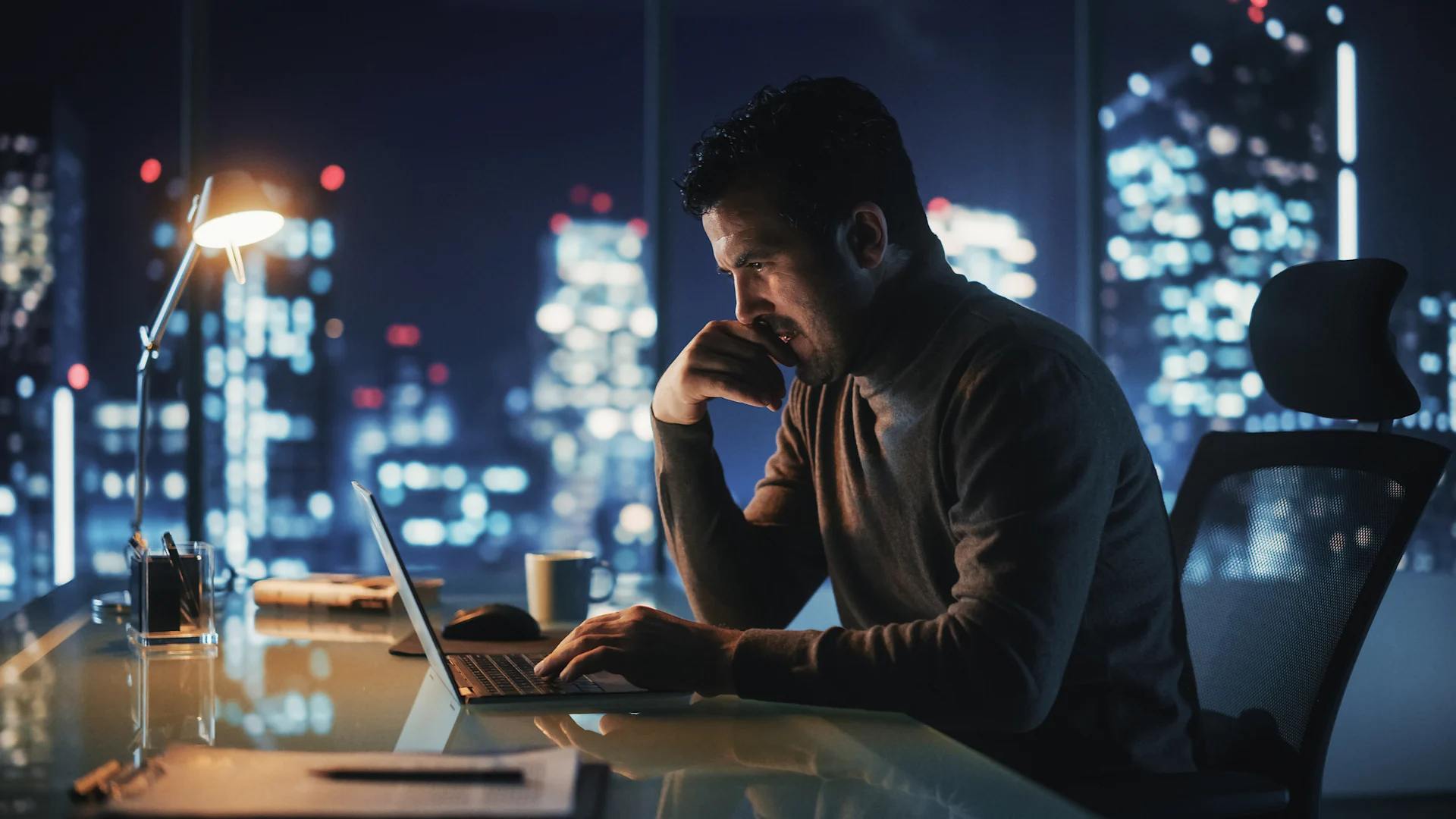 A man at a desk using a laptop, with city lights twinkling in the background, highlighting a dynamic urban environment.
